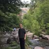 A person standing by a rocky river surrounded by lush greenery.